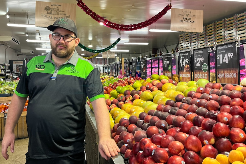 Man in black and green polo shirt wearing glasses and a hat stands next to a fruit display in a grocer. 