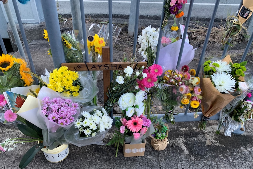 Flowers lay beside a wooden cross by the side of the road. The cross has "RIP Aysha" on it.