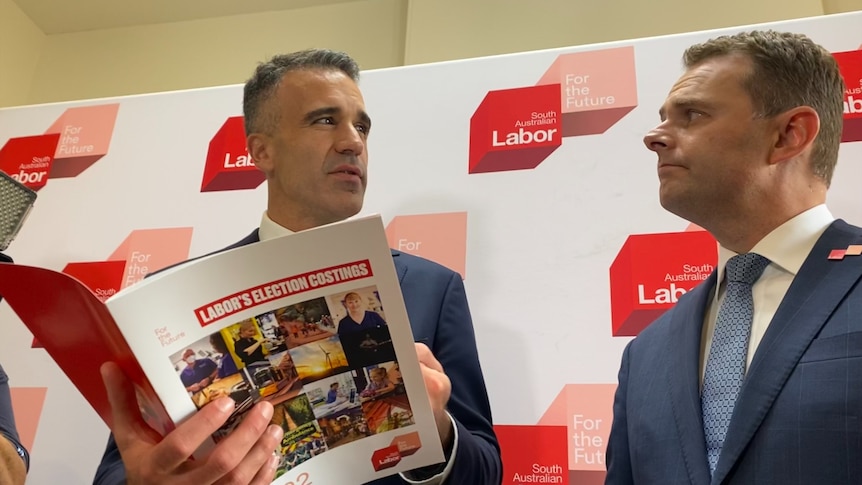 A man holding a brochure titled 'Labor's election costings' and another man look at each other in front of a Labor backdrop