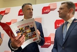 A man holding a brochure titled 'Labor's election costings' and another man look at each other in front of a Labor backdrop