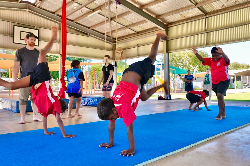 Four kids perform handstands on a blue circus mat, wear red tee, black shorts, man watches, rattan roof, open on all sides.