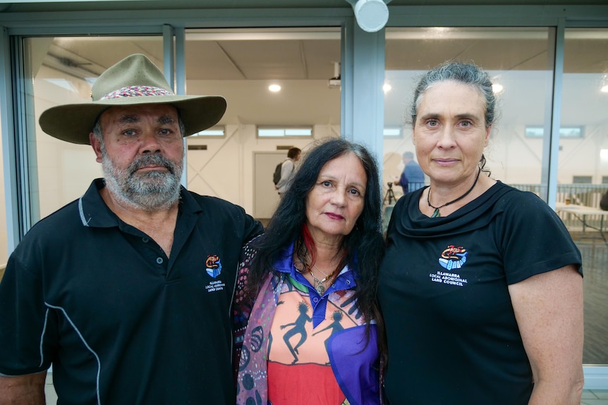 A man and two woman, all middle-aged and Indigenous, stand with their arms around each other, looking solemn.