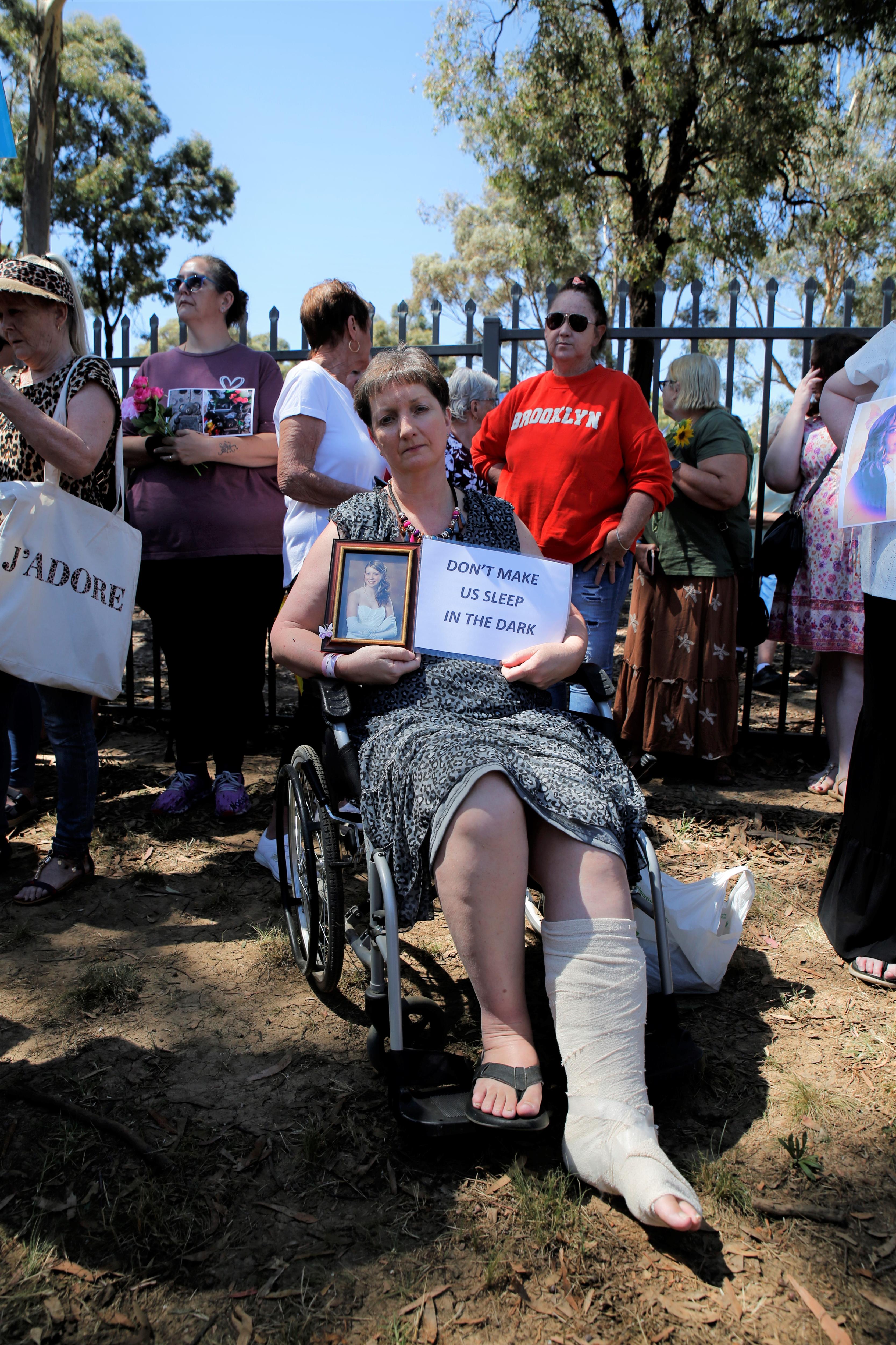 A woman in a wheelchair outside the cemetery, holding a photo and a sign.