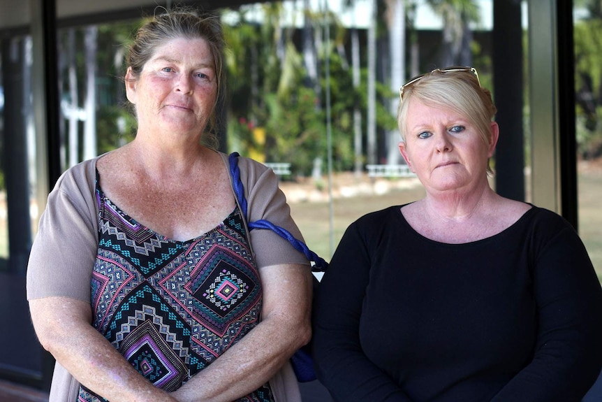 Two women stand outside a building with reflective glass, they have serious expressions.