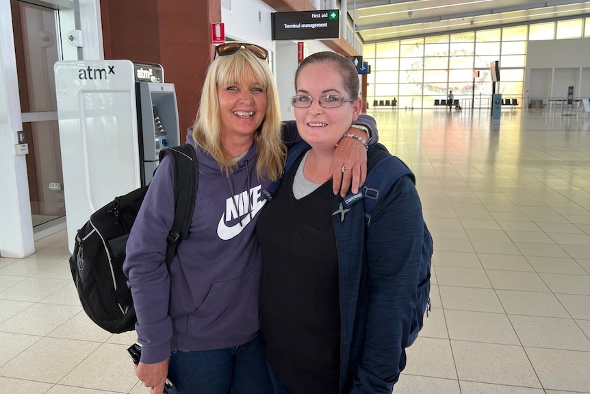 Two woman stand smiling and posing for a photo in an airport terminal, with their hands around each other's shoulders.