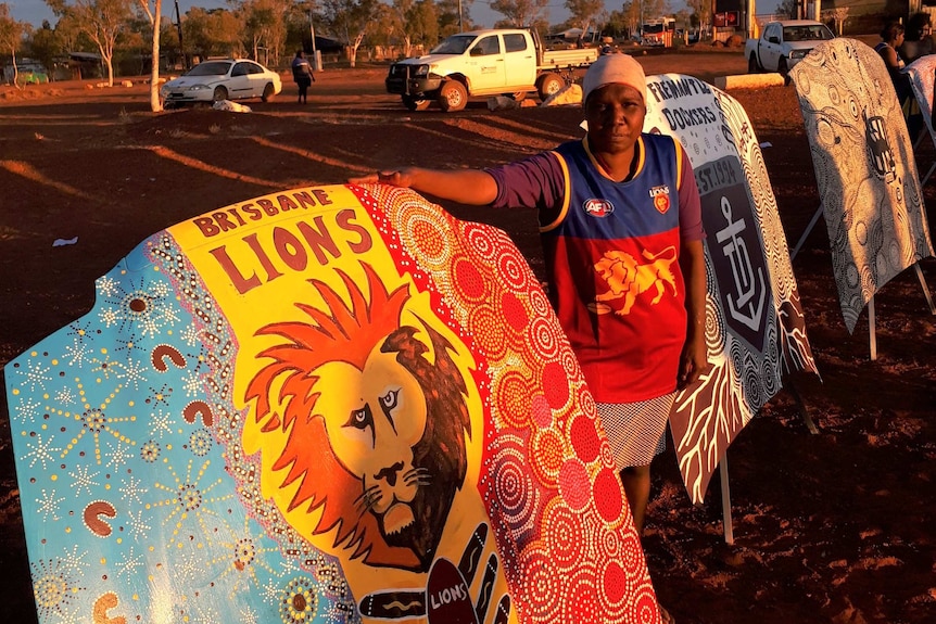 An Indigenous man wearing a football jersey stands next to a bonnet emblazoned with the Brisbane Lions logo.