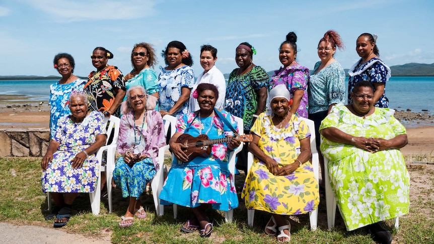 14 women wearing colourful dresses sit on the shore of a beach. One holds a ukulele. All are smiling.