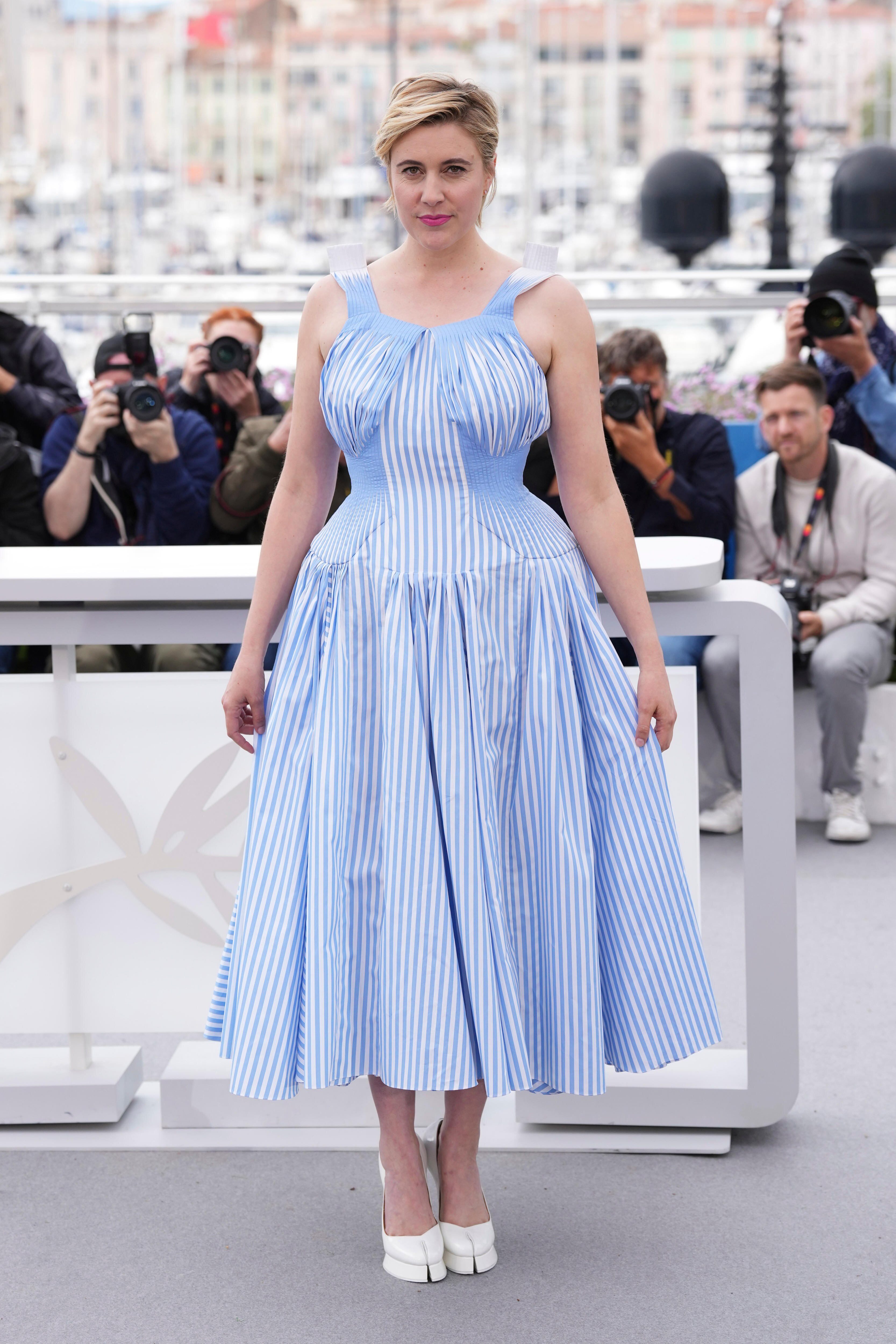 Greta Gerwig wearing a blue and white striped dress with corset-like detailing and a pair of white split-toed heels