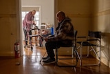 An elderly woman with a walker stands in the kitchen while an older man sits in a chair in the foreground.