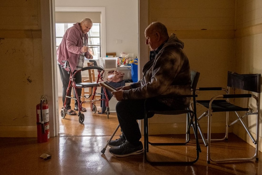An elderly woman with a walker stands in the kitchen while an older man sits in a chair in the foreground.