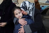 A girl with scabs and bruises on her forehead is hugged by a women in a black and white headscarf