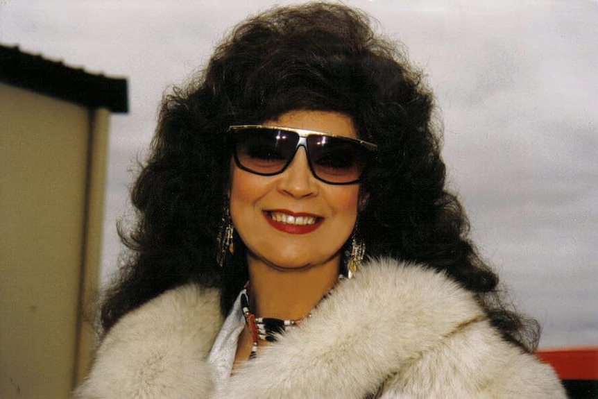 a woman with big hair wearing sunglasses looks at the camera smiling 