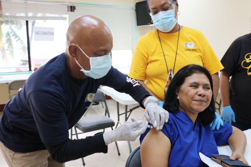 A woman with black hair and an electric blue blouse sits at a vaccination centre and receives a needle jab in her arm.