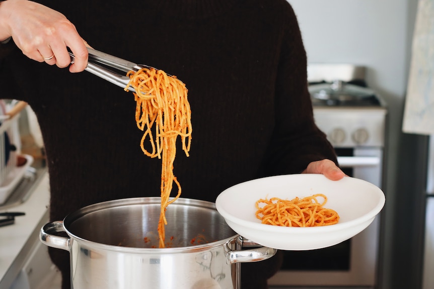 Using tongs to serve freshly made red pesto pasta into a bowl, a vegetarian dinner.