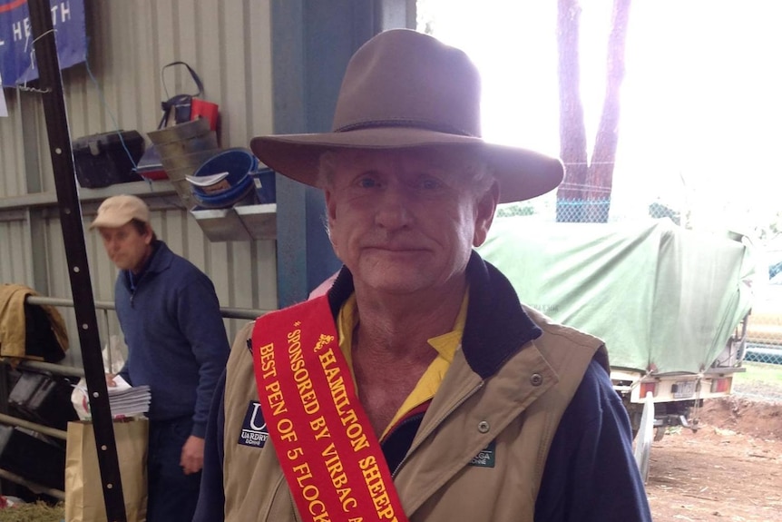 An elderly man wearing a hat and a sash stands in a shed.