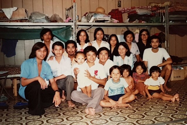 A group of 18 Vietnamese refugees, including women, men and children, sit on a linoleum floor in front of six bunk beds in 1981.