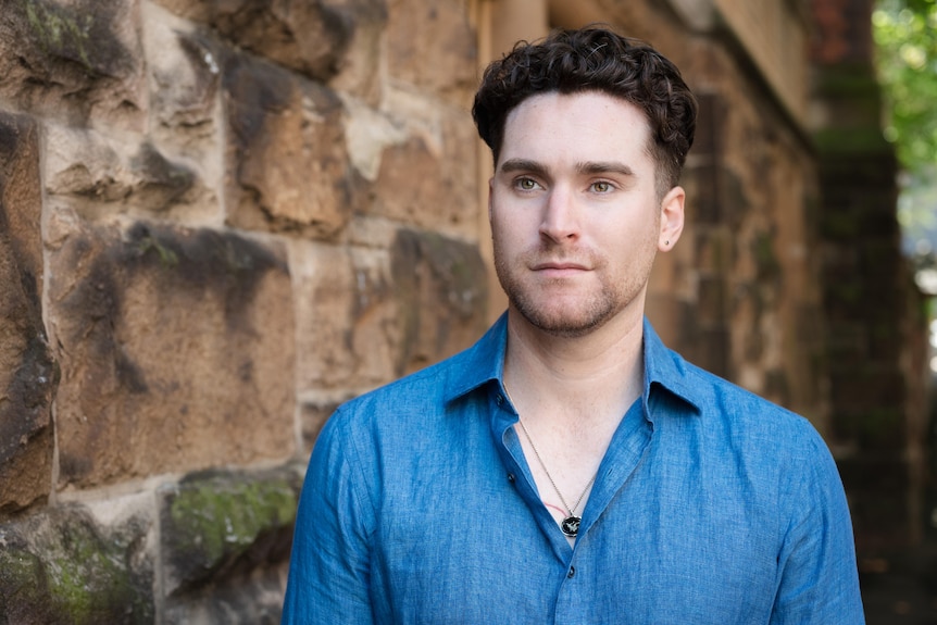 A young man stands in front of a rough brick wall, he is looking past the camera with a neutral expression.