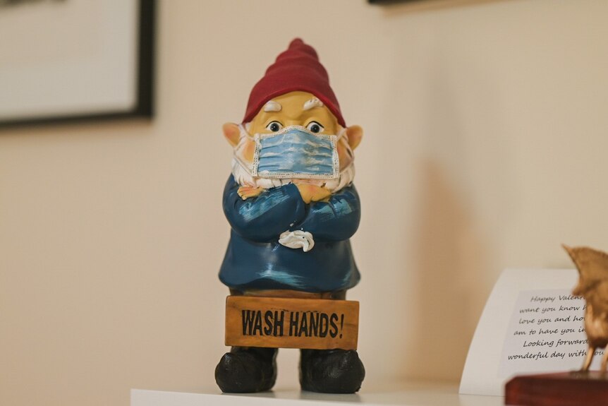 A garden lgnome wearing a medical mask witha  sign reading WASH YOUR HANDS. 