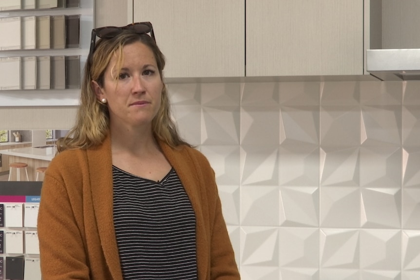 A woman with blond hair standing in front of a white tile splashback stares at a camera