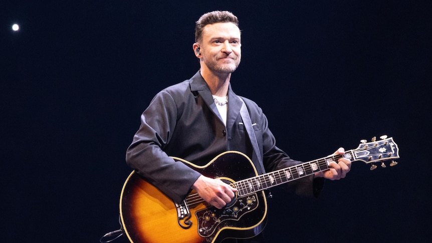 A man stands smiling on stage, strumming a guitar around his neck. He wears a grey button up shirt.