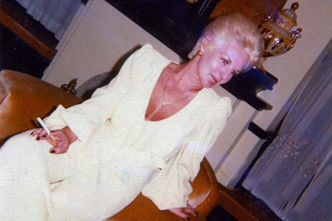 Shirley Finn, in a cream formal jumpsuit, leans back on an arm chair, holding a cigarette in one hand. Her nails are painted red