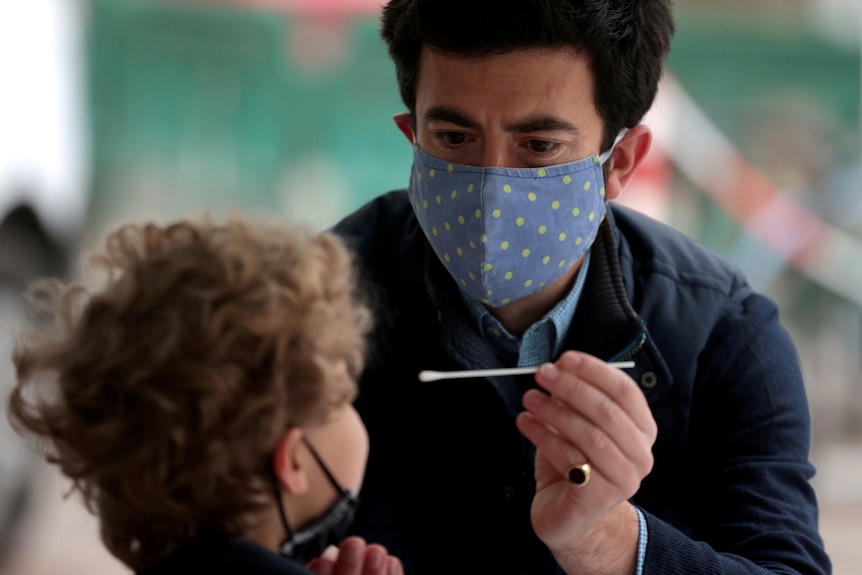A man in a mask holds a swab in front of a child's face 