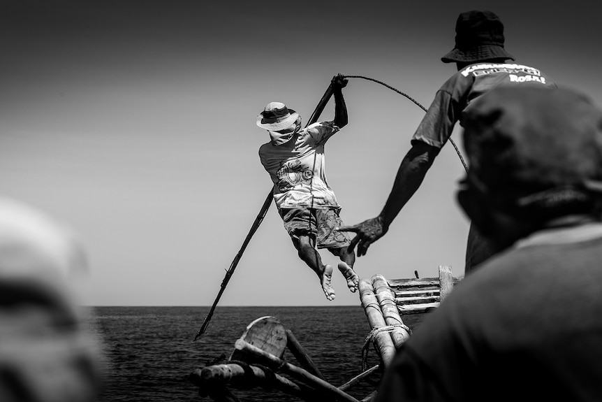 A black and white photo of a man diving off a boat with a spear.
