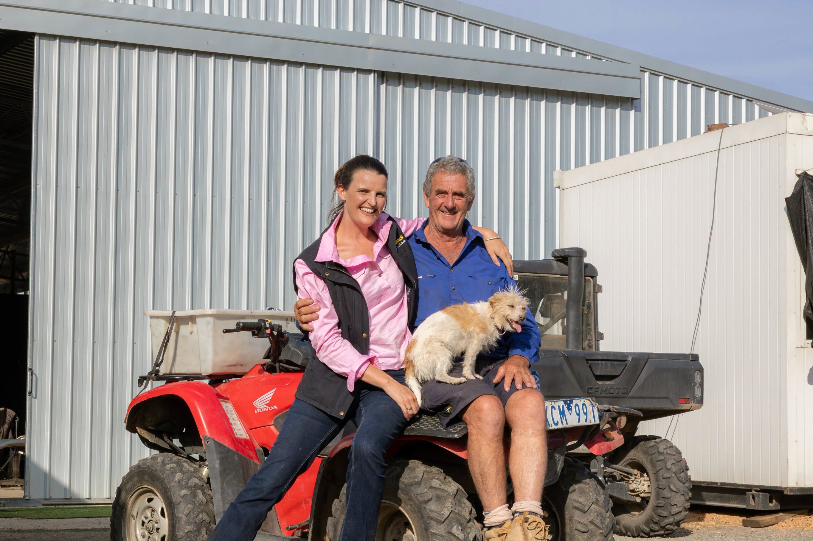 Annabelle Cleeland sits with her arms around a man's shoulder on the back of an ATV on a farm.