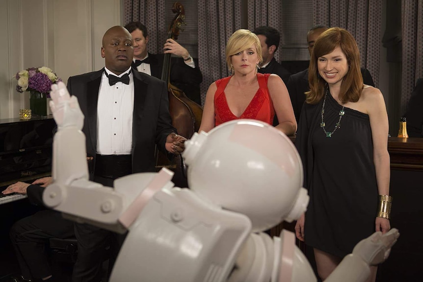 The three actors stand reacting in front of a robot