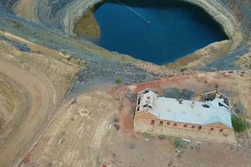 Birds-eye view of derelict building with some roofing missing and beams exposed next to edge of open-cut mine