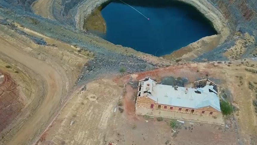 Birds-eye view of derelict building with some roofing missing and beams exposed next to edge of open-cut mine