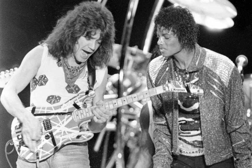 Michael Jackson, on stage, watches on as Eddie Van Halen plays the guitar next to him, both with their mouths open