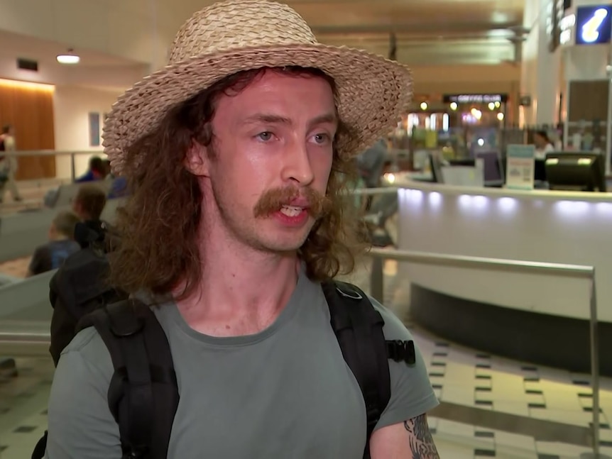 A young man with a moustache, long hair and a straw hat speaks at an airport.