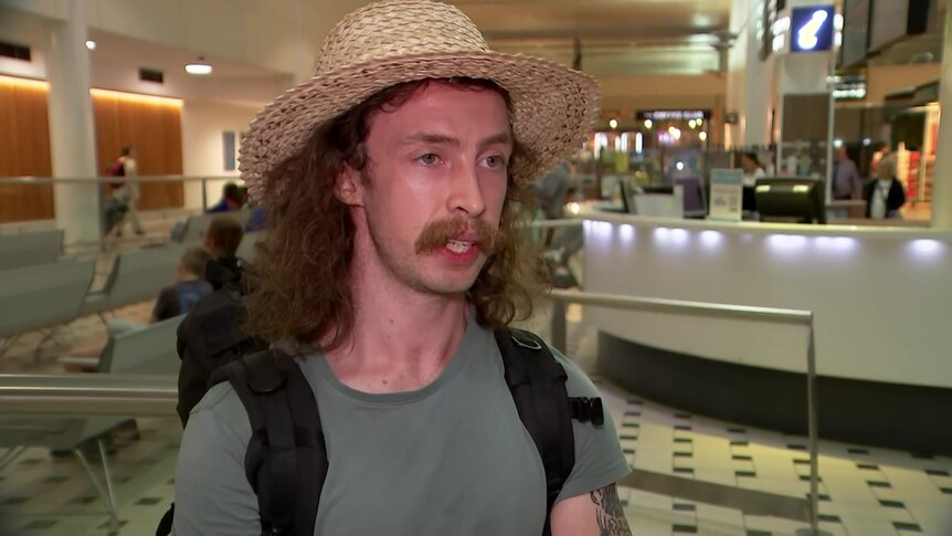 A young man with a moustache, long hair and a straw hat speaks at an airport.
