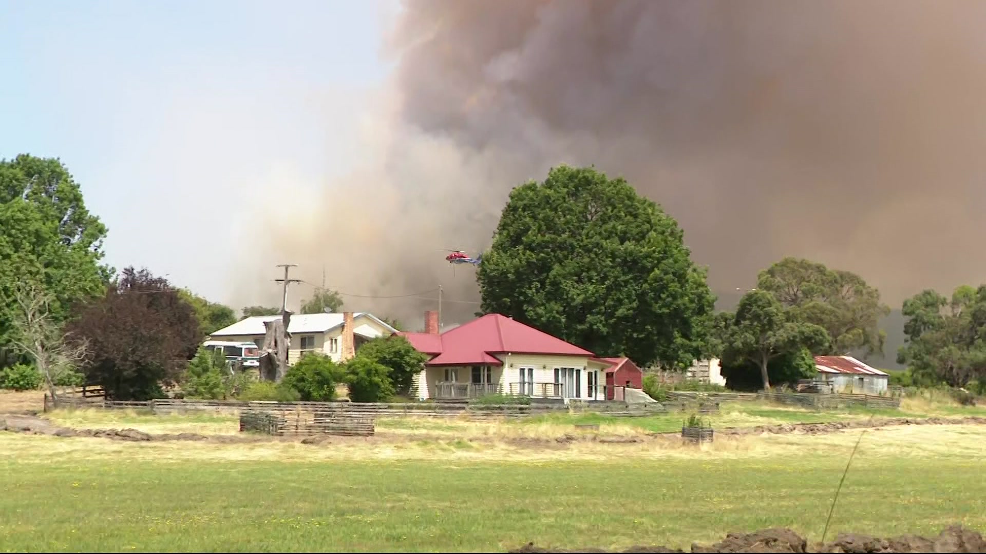 A helicopter flies through thick dark smoke that billows into the sky behind a wooden house with a red roof.