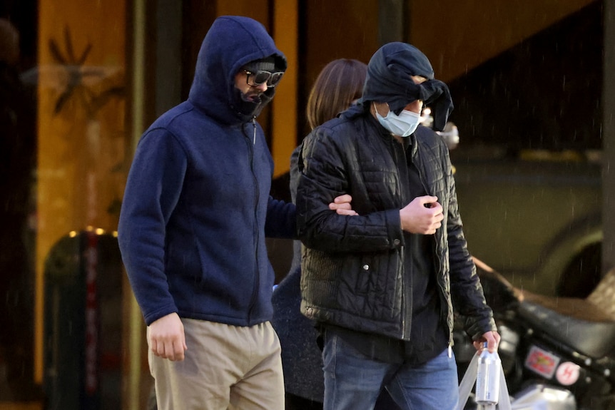 A man wearing sunglasses and a blue hoodie pulled tighly over his face walks out of a building holding someone's arm.