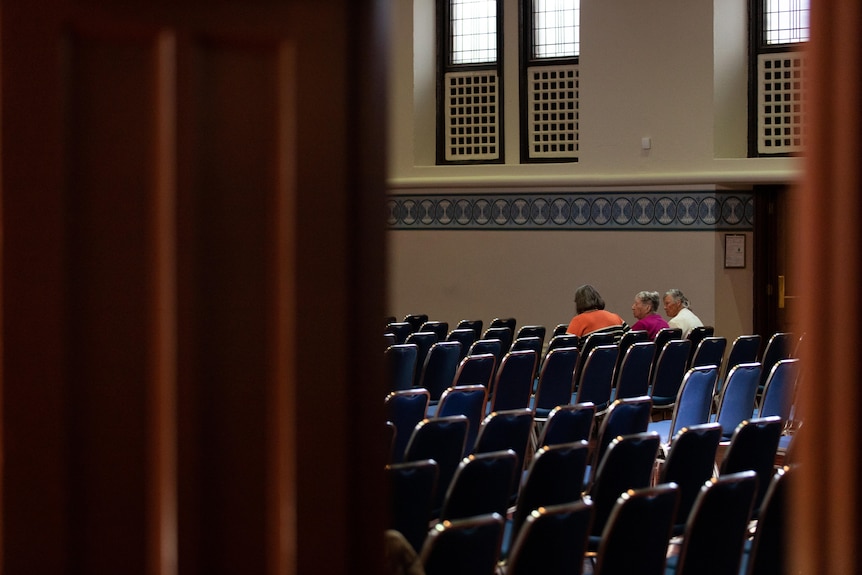 Three people sit in a large room full of rows of seats.