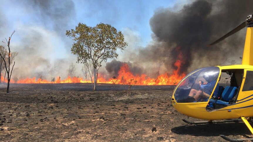 Flames from a bushfire rise into the air with a yellow helicopter off to the right of the shot.
