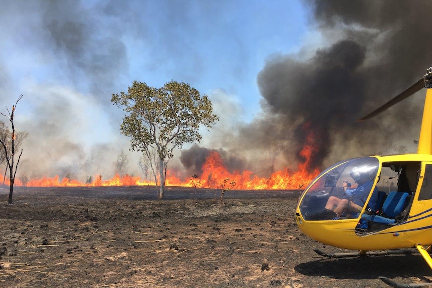 Flames from a bushfire rise into the air with a yellow helicopter off to the right of the shot.