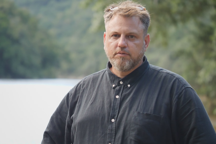 Fair-haired man with beard stands in front of lake with concerned expression and sunglasses pushed up on his head