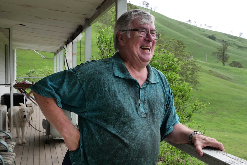 An older man stands on a wooden deck, laughing and looking out at green hills.