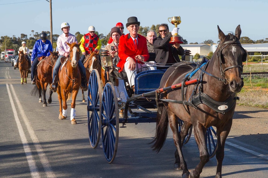 A horse and cart with riders on horse back walking in a country town street with a man holding up a gold trophy