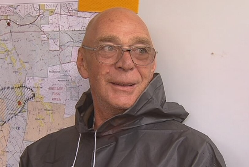 A bald man wearing glasses and a rain poncho stands in front of a map on a pin-up board.