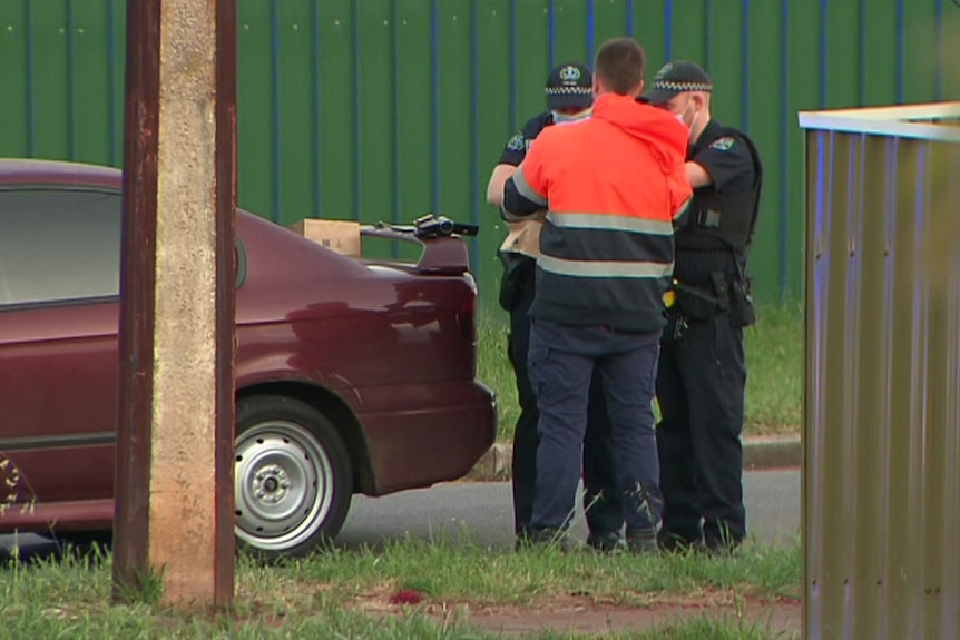 A man wearing an orange and blue jumper is is arrested by police next to a car and metal fence
