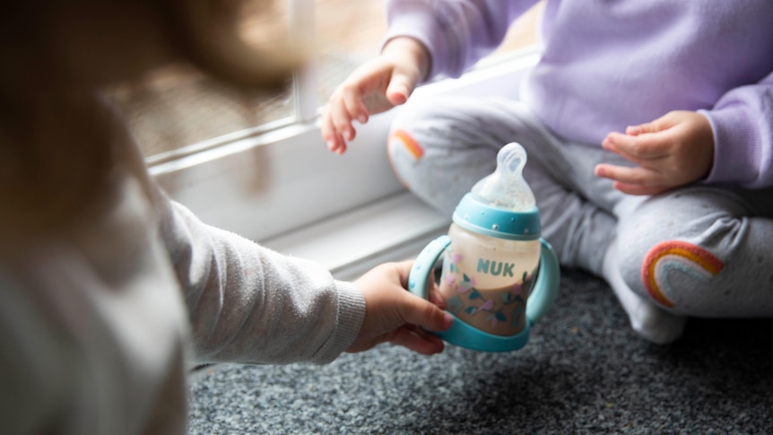 A child hands a bottle of baby formula milk to a two-year-old.