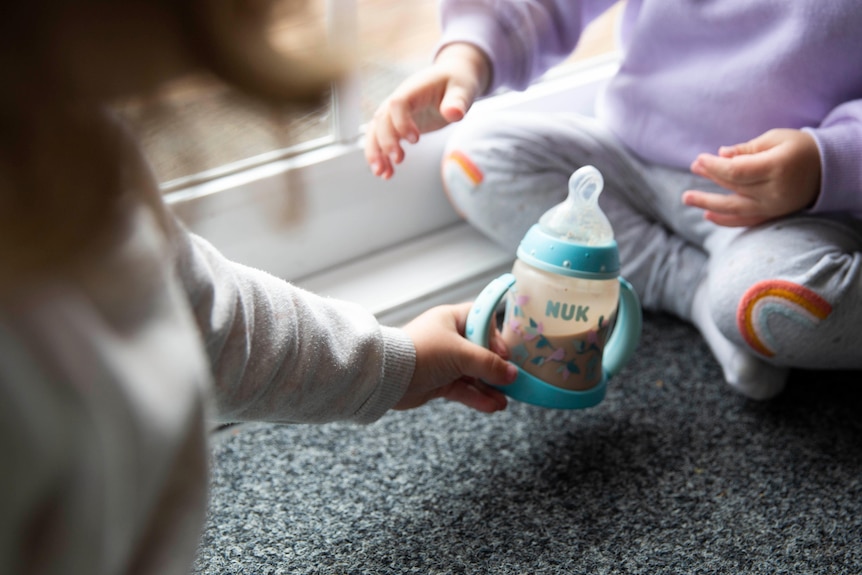 A child hands a bottle of baby formula milk to a two-year-old.