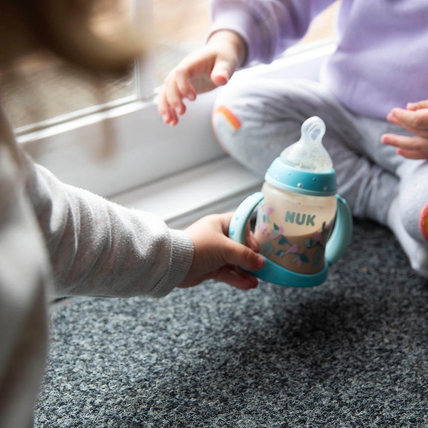 A child hands a bottle of baby formula milk to a two-year-old.