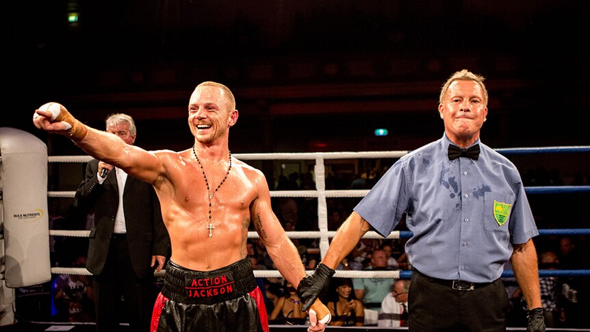 A man wearing red and black boxing shorts pointing to the crowd and smiling in a boxing ring.