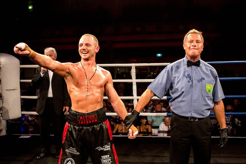 A man wearing red and black boxing shorts pointing to the crowd and smiling in a boxing ring.