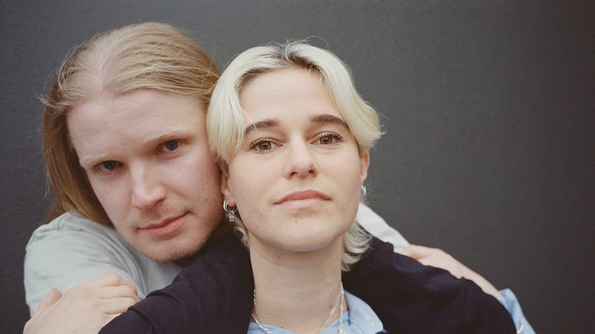 Closeup of two people with light hair smiling slightly at the camera. Their heads are next to each other.